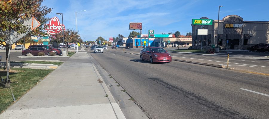 View looking down a busy commercial street with cars driving in both directions. A sidewalk runs alongside the road with fall-colored trees. Visible businesses include Arby’s on the left and Subway on the right. The sky is clear and blue.