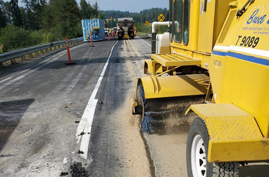 A broomer follows a truck putting down sand