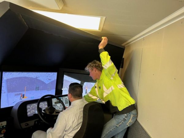 A person wearing a high-visibility yellow jacket leans over the shoulder of another person seated at a driving simulator. The simulator screens display a snowy road scene as the standing person provides instruction or guidance.
