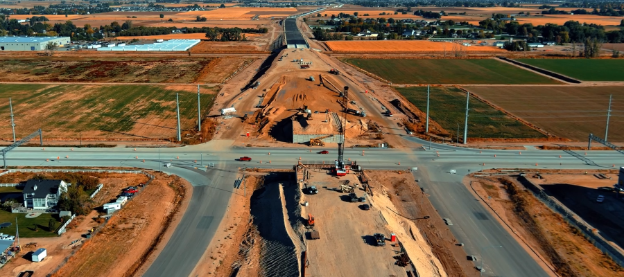 Aerial view of a large highway construction project cutting through open farmland. The image shows a major intersection where crews are building an overpass or interchange, with heavy equipment, cranes, and dirt embankments visible on both sides. Traffic cones line the crossroad, and nearby fields and a few buildings stretch into the distance under a clear blue sky.